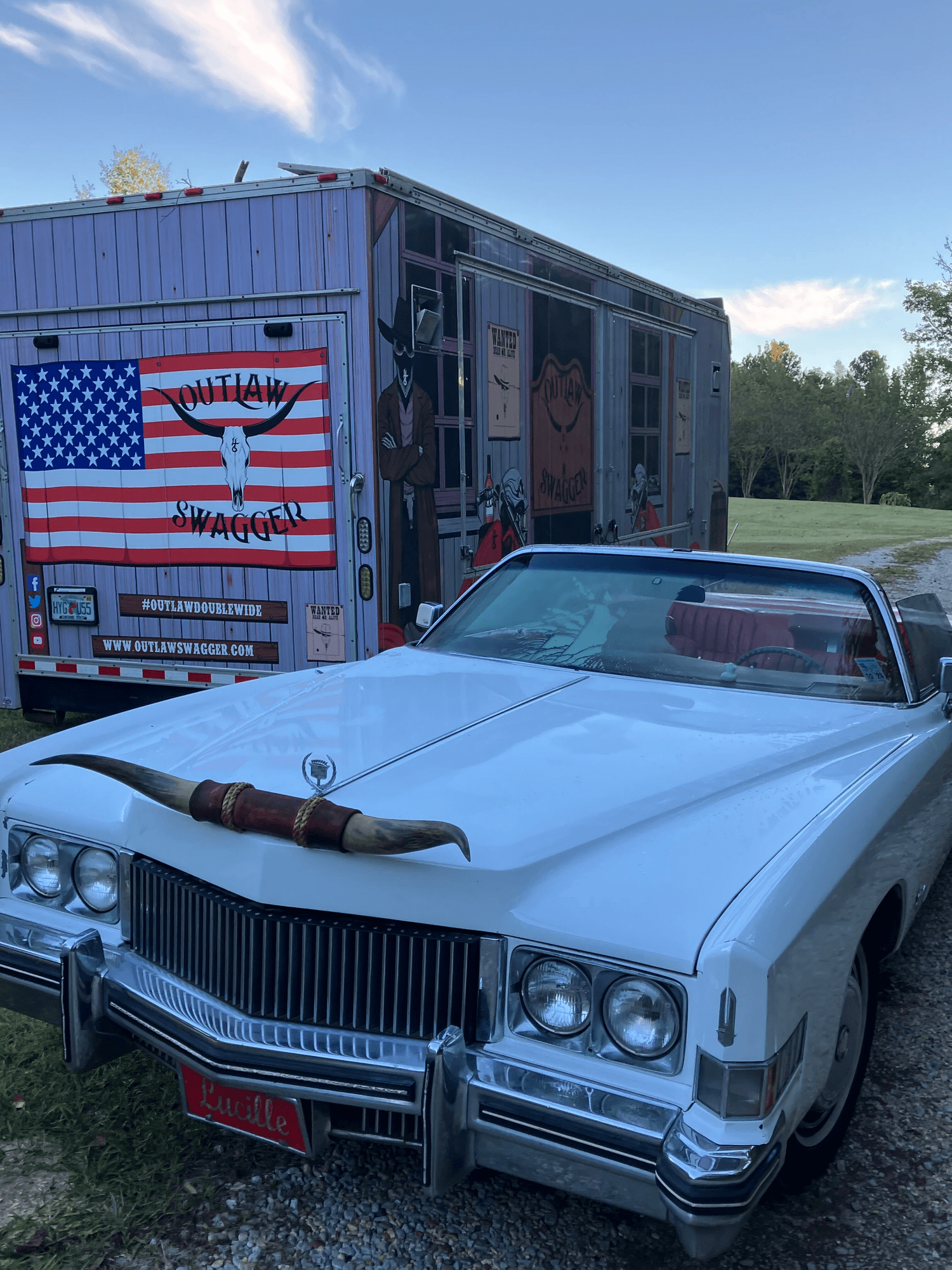 A white Cadillac with longhorns on the hood parked beside the Outlaw Swagger trailer